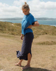 Person wearing a blue top and navy skirt standing on a grassy hill with a scenic background.