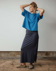 Woman wearing a blue top and dark skirt standing against a plain wall.