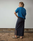 Woman wearing a blue top and dark skirt standing against a plain wall with wooden floor.