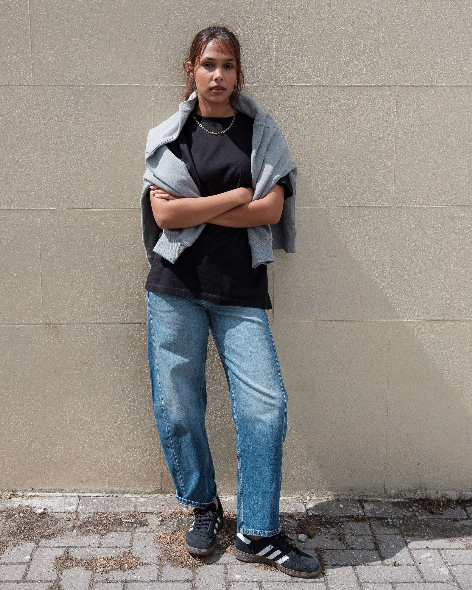 Person wearing a black shirt, blue jeans, and a gray sweater against a beige wall.
