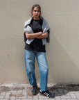 Person wearing a black shirt, blue jeans, and a gray sweater against a beige wall.