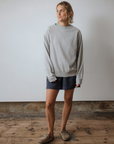 Person wearing a grey sweatshirt and dark shorts standing on a wooden floor with a white wall background