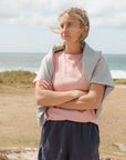 Woman standing on a beach with arms crossed, wearing a pink shirt and grey jumper.
