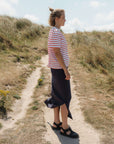 Woman in a striped shirt and dark skirt standing on a sandy path with grass and flowers around.