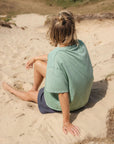 Person sitting on a sandy beach wearing a green shirt and dark shorts.