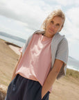 Woman standing on a sandy beach wearing a pink top and grey sweater.