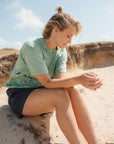Woman sitting on a sand dune wearing a light green t-shirt and dark shorts.
