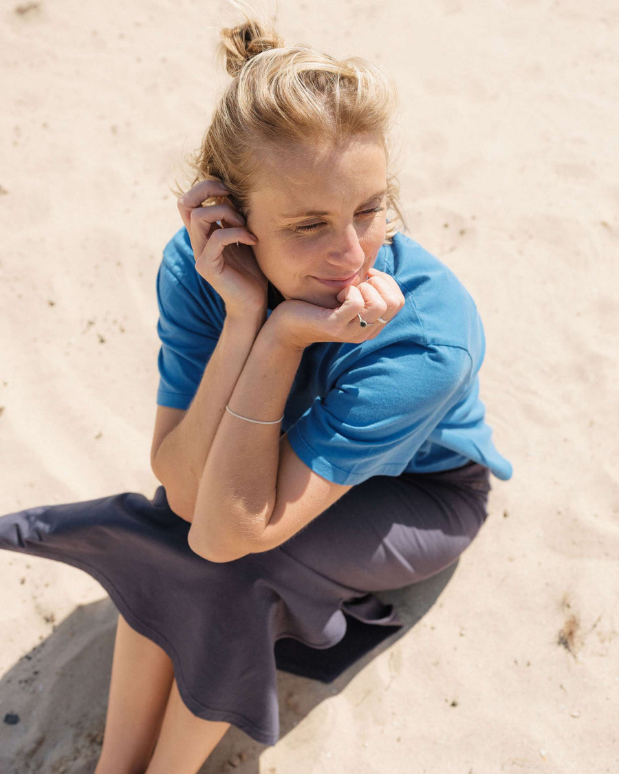 Woman sitting on a sandy beach wearing a blue shirt and dark pants.