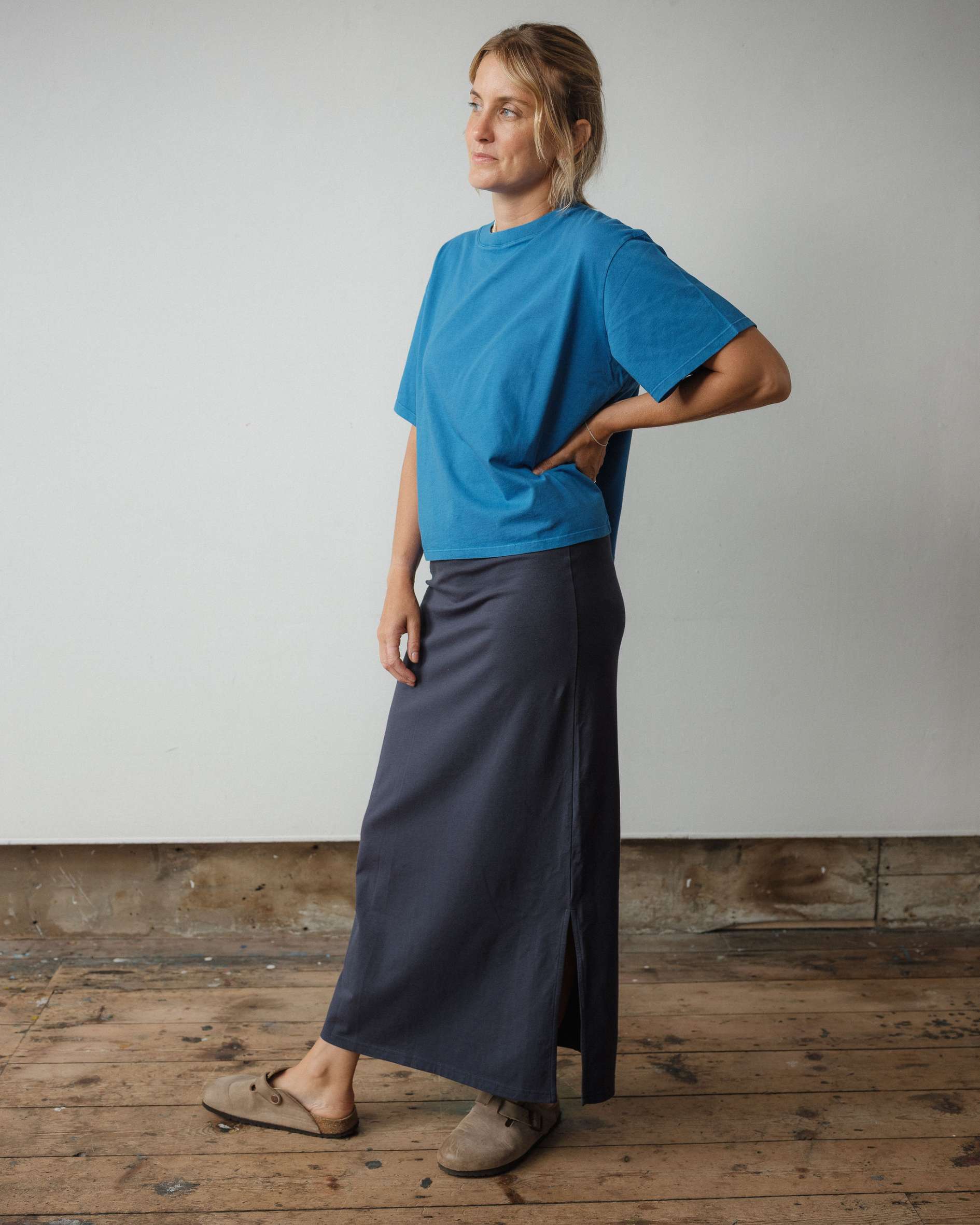 Person wearing a blue shirt and dark skirt standing on a wooden floor with a plain wall background