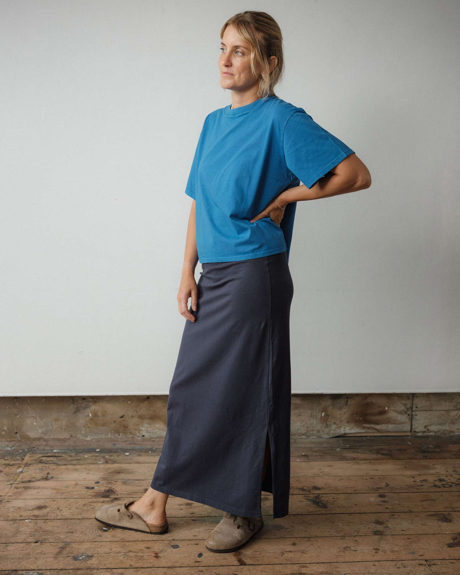 Person wearing a blue shirt and dark skirt standing on a wooden floor with a plain wall background