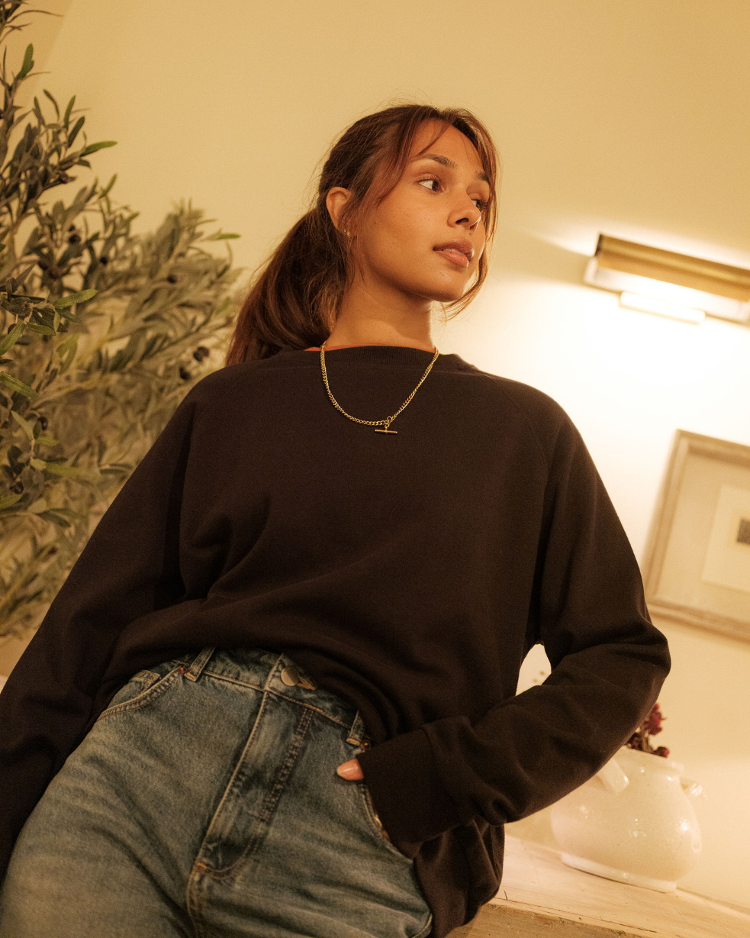 Woman wearing a black sweater and blue jeans indoors with a plant in the background