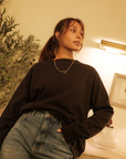 Woman wearing a black sweater and blue jeans indoors with a plant in the background