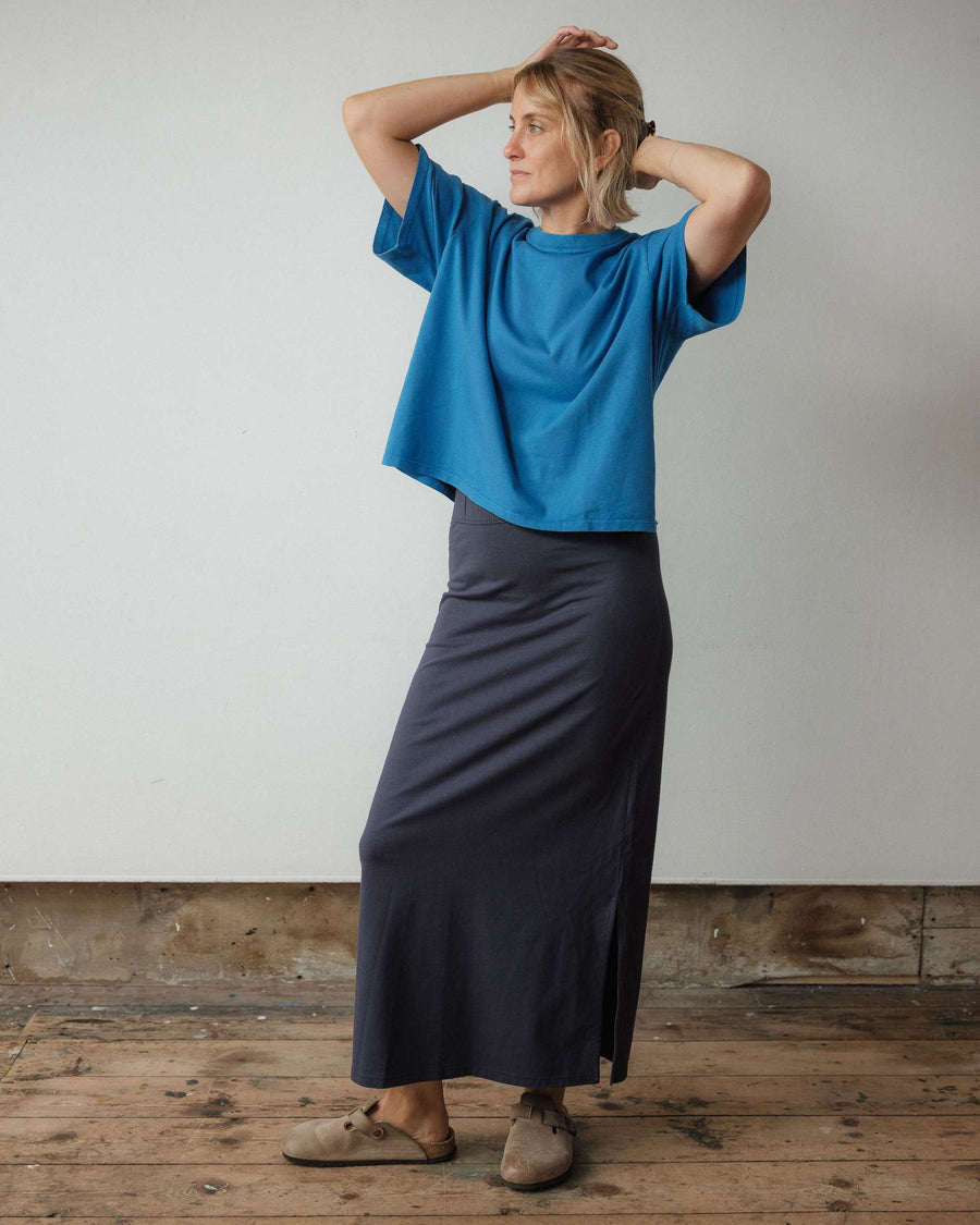 Woman wearing a blue top and dark skirt standing against a plain wall.