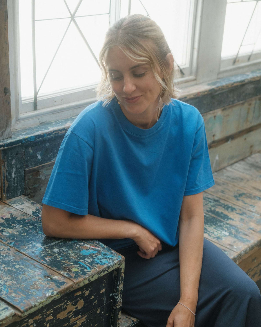 Person wearing a blue shirt sitting on a rustic wooden bench with large windows in the background.