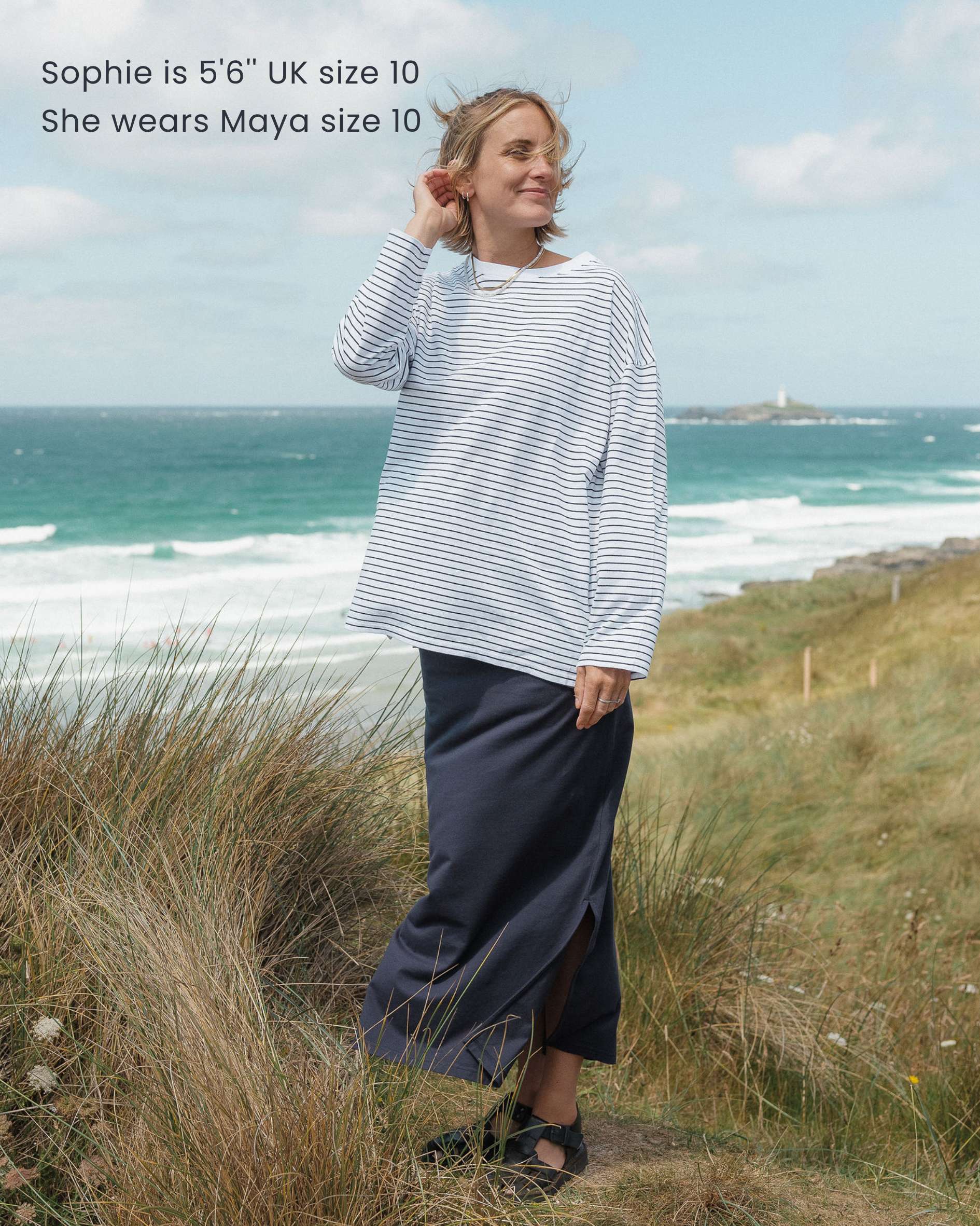 Woman standing on a grassy hill by the ocean wearing a striped t-shirt and dark skirt.