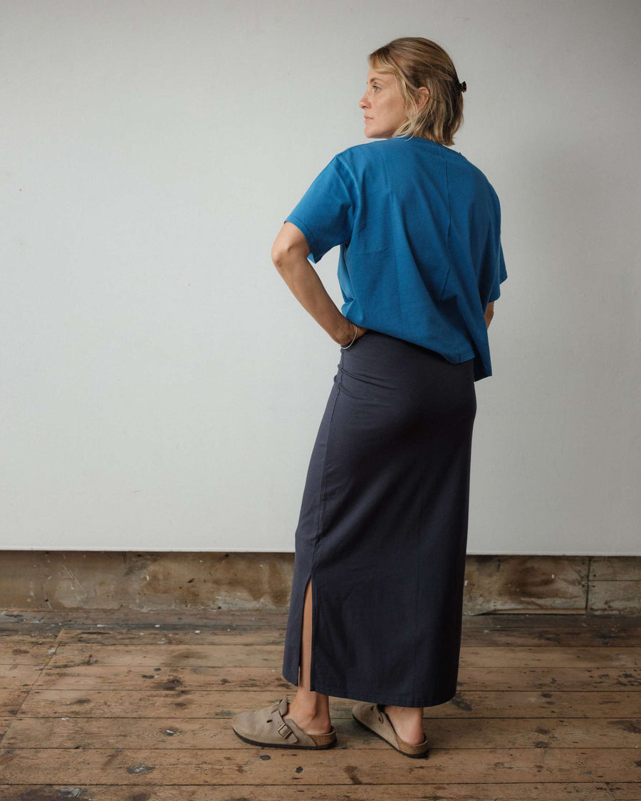Woman wearing a blue top and dark skirt standing against a plain wall with wooden floor.