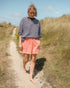Woman in a striped shirt and pink shorts standing on a sandy path with grass and blue sky in the background