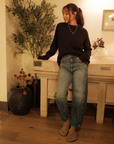 Woman in black sweater and blue jeans standing indoors with decorative plants and furniture.