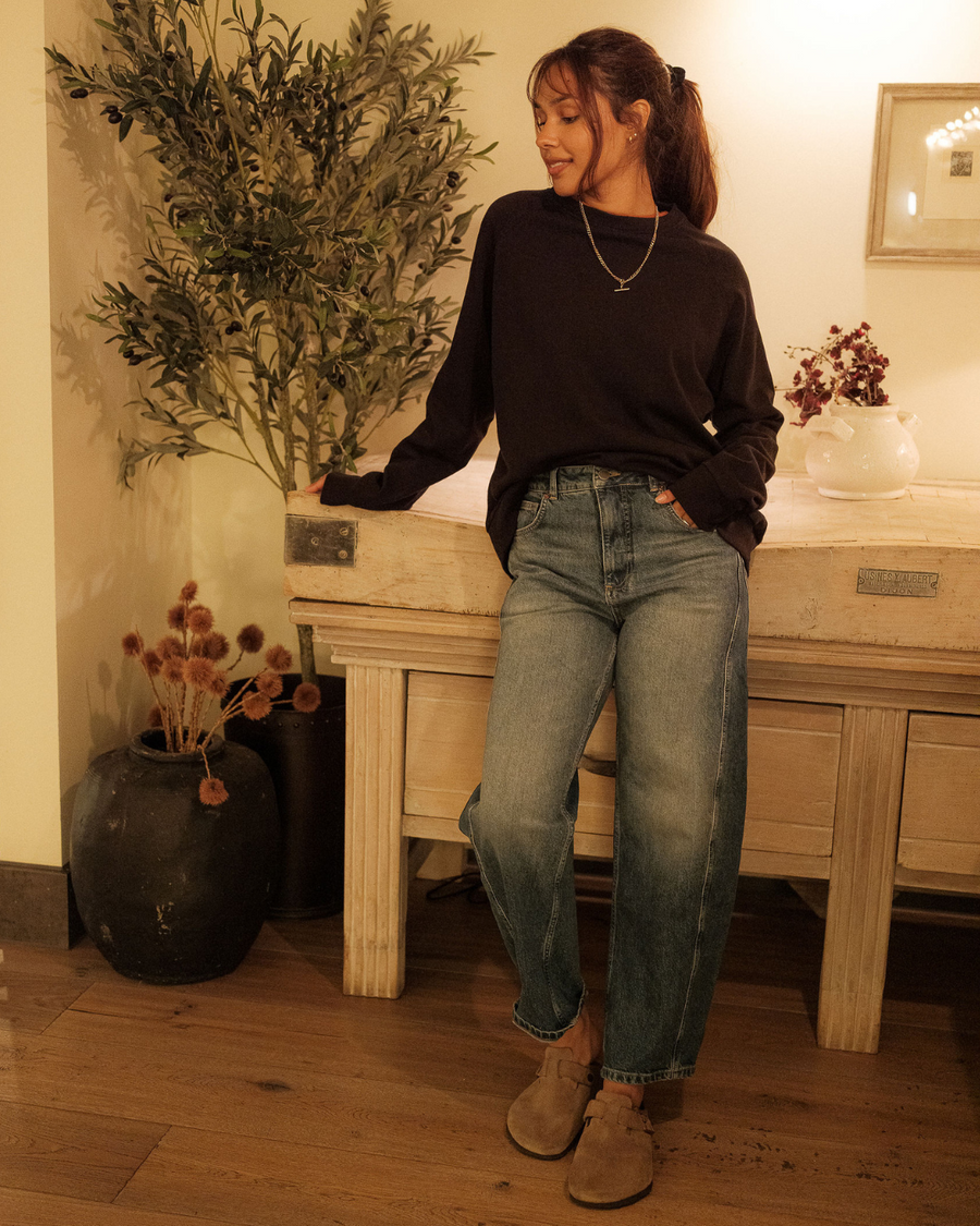 Woman in black sweater and blue jeans standing indoors with decorative plants and furniture.