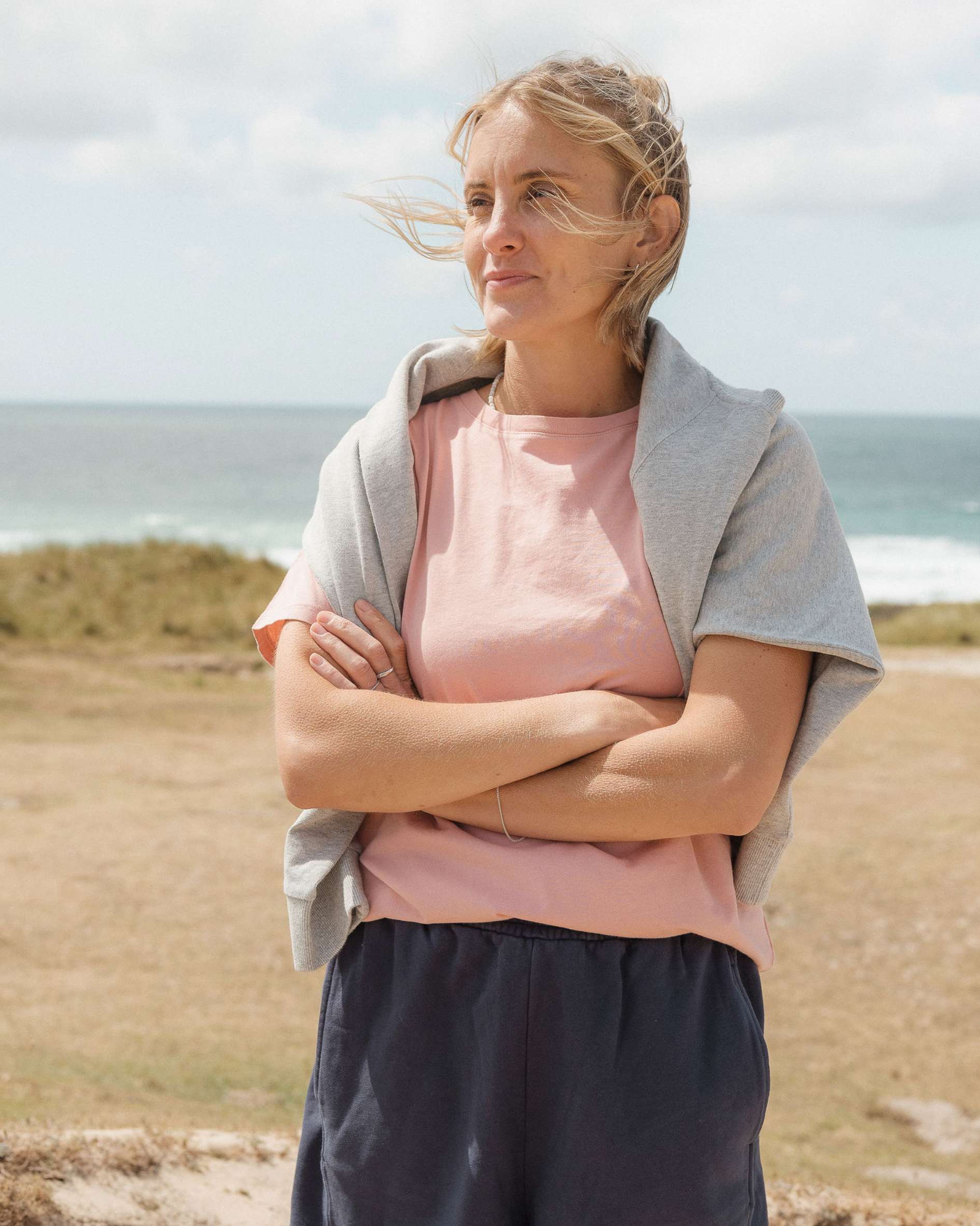 Woman standing on a beach with arms crossed, wearing a pink shirt and grey jumper.