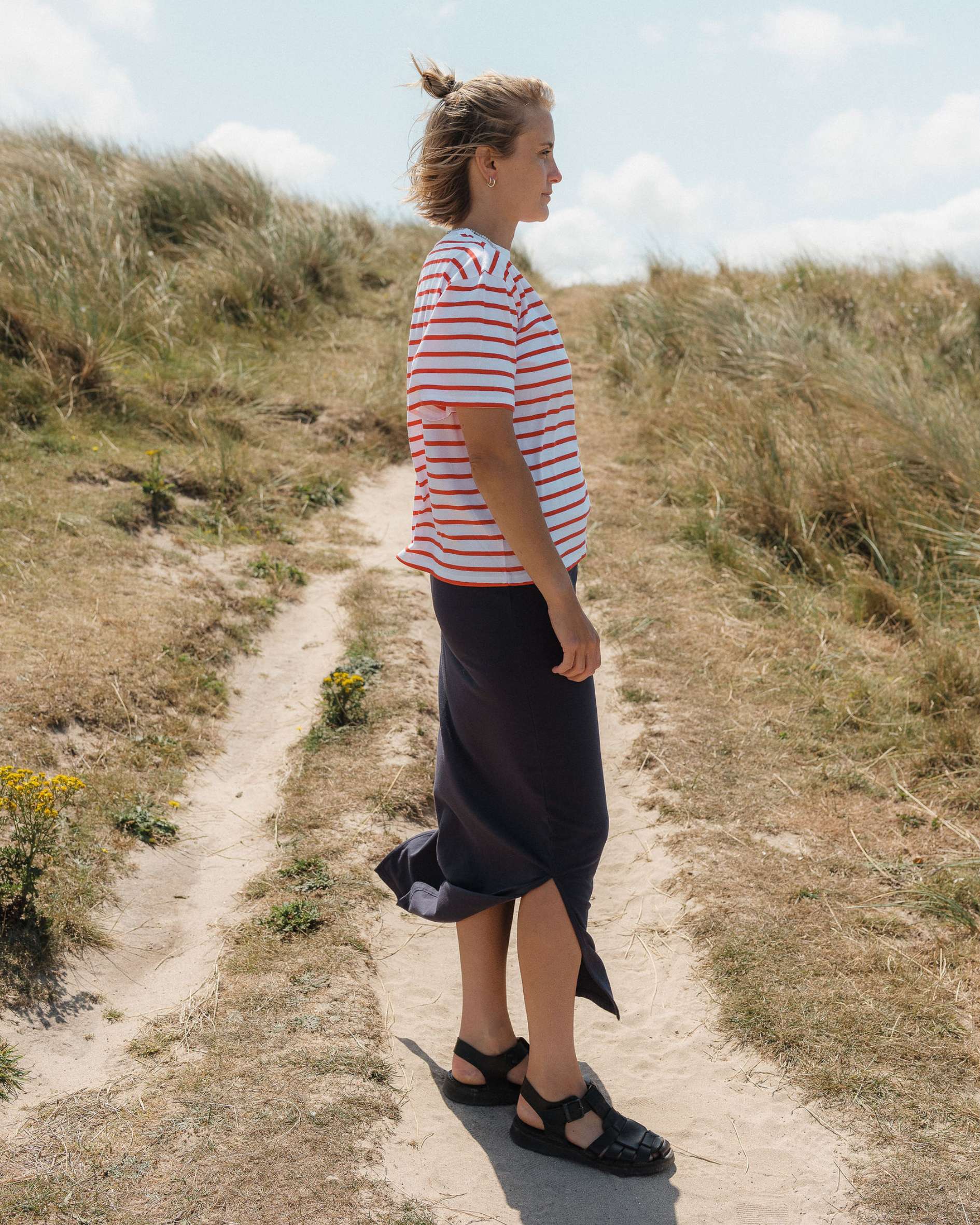 Woman in a striped shirt and dark skirt standing on a sandy path with grass and flowers around.