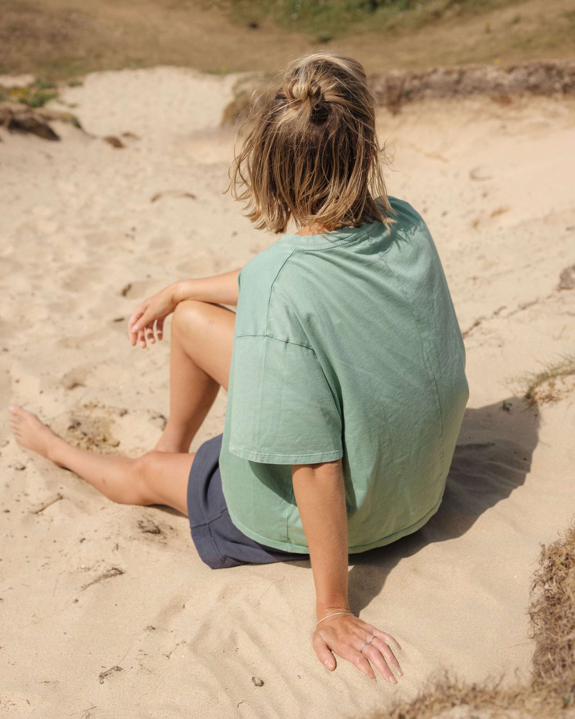 Person sitting on a sandy beach wearing a green shirt and dark shorts.