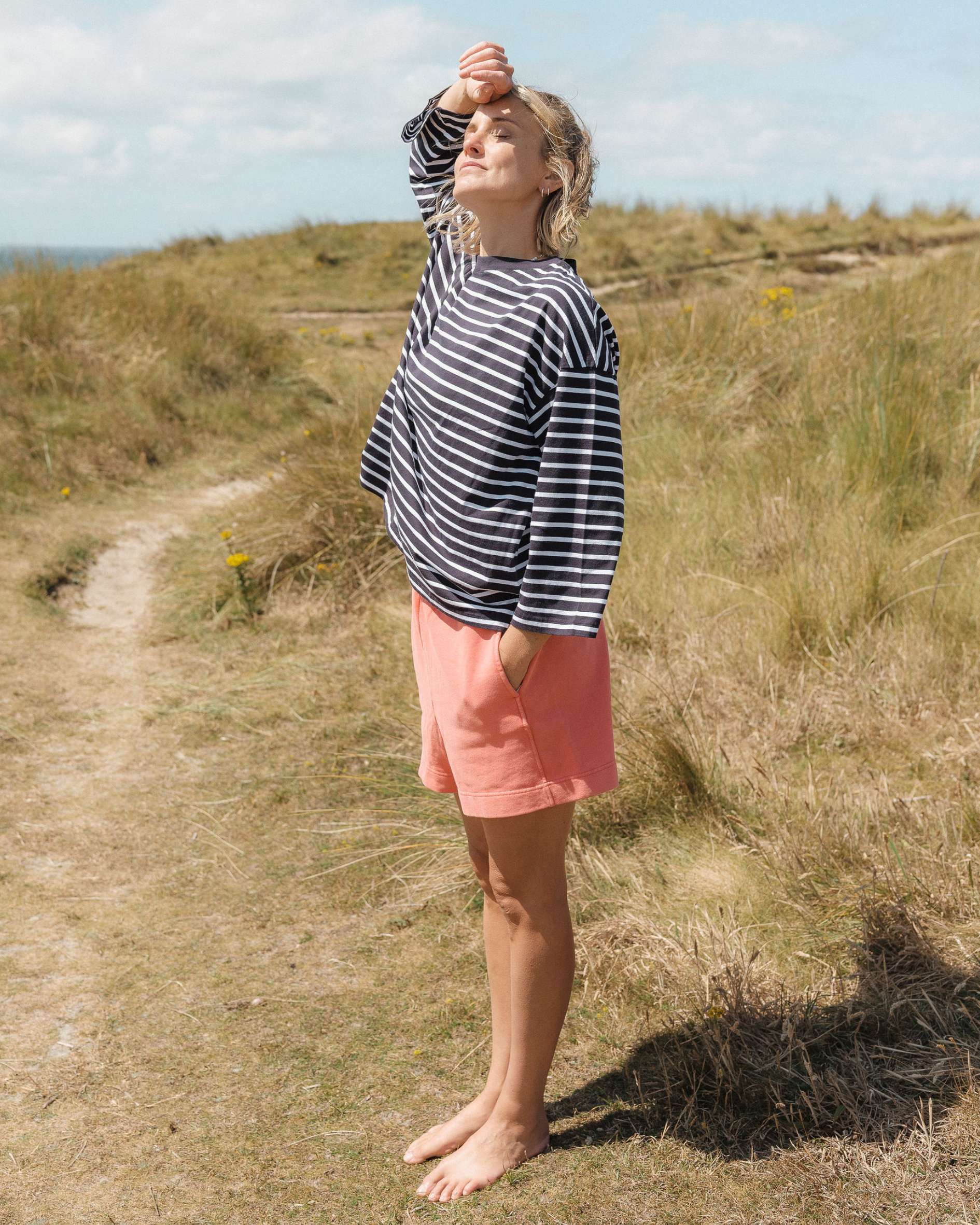 Woman in a Breton striped tee shirt and pink shorts standing on a grassy path with a clear sky.