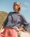 Woman wearing a navy blue and white t-striped shirt with pink sweat shorts standing outdoors against a blue sky.