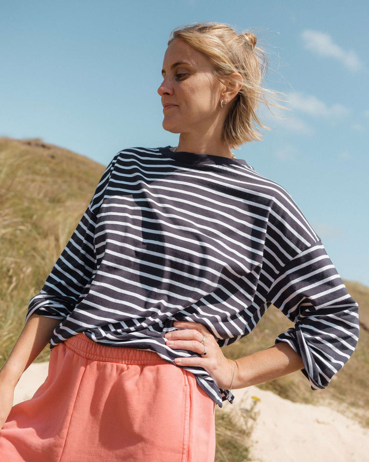 Woman wearing a navy blue and white t-striped shirt with pink sweat shorts standing outdoors against a blue sky.